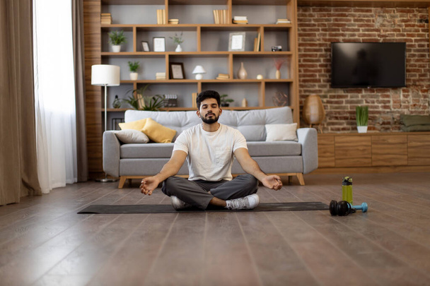 Healthy young indian man doing workout at home with meditation exercise. Sporty bearded guy sitting on lotus position with mudra gesture and looking at camera. - Foto, Imagem