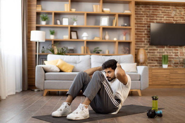 Active indian man in sport clothes lying on yoga mat and training abs with body crunches at home. Fitness workout of strong young guy. Healthy lifestyles concept. - Foto, imagen