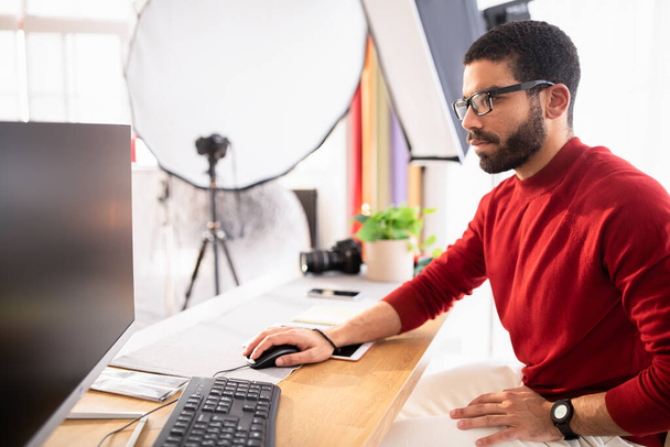 Όμορφος γενειοφόρος φωτογράφος της χιλιετίας από τη Μέση Ανατολή φορώντας γυαλιά καθισμένος στο τραπέζι, δουλεύοντας σε υπολογιστή με μαύρη άδεια οθόνη στο στούντιο του γραφείου, ρετουσάρισμα φωτογραφιών, πλαϊνή άποψη - Φωτογραφία, εικόνα