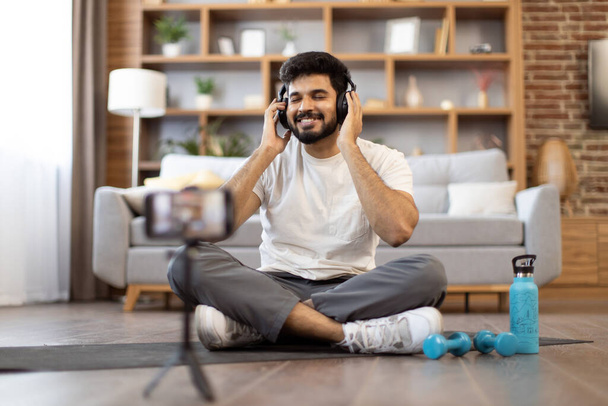 Attractive arabic man in sports clothes enjoying music in headphones while sitting cross-legged on floor. Happy instructor shooting video on smartphone about using headset for gym while working out. - 写真・画像