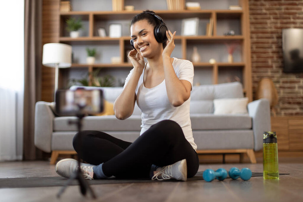 Pretty multiracial vlogger in headphones smiling at phone camera on tripod while sitting on yoga mat near dumbbells and waterbottle. Active woman recording workout video for sport vlog in apartment. - Fotoğraf, Görsel