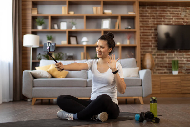 Cheerful blogger greeting her followers at online training exercise held via mobile phone on tripod in cozy sitting room. Active multiracial woman motivating participants to lead healthy lifestyle. - Photo, Image