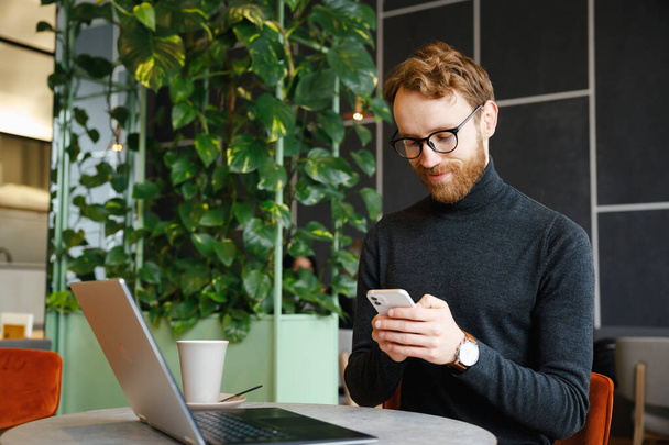 A young red-haired guy, a programmer or businessman in glasses, sits in a stylish cafe at a laptop and chats in a messenger on a smartphone, checks mail. Freelancer works remotely. - Foto, Imagen