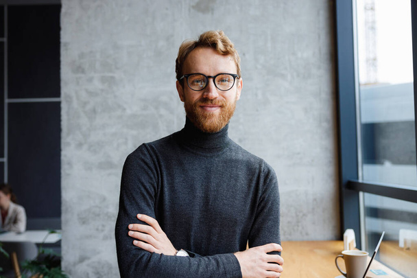 A young red-haired guy, a programmer or an entrepreneur in glasses, in a stylish cafe Looks at the camera and smiles. Freelancer works remotely. Online communication. small business - Foto, afbeelding