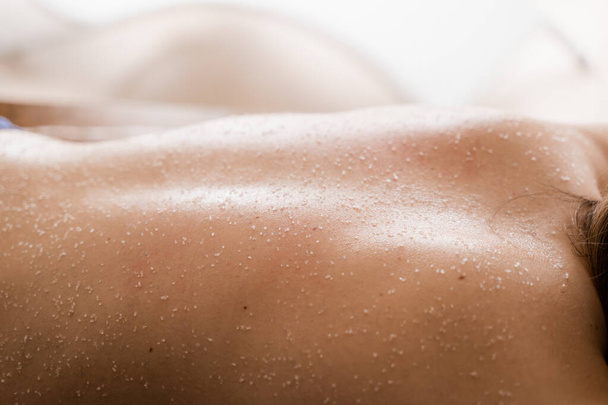 Соль скраб на спине девушки в спа-салоне крупным планом. Косметологическая процедура очистки, пилинга для омоложения кожи - Фото, изображение