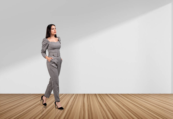 Thoughtful businesswoman walking with hands in pocket on hardwood floor, white copy space blank background. Concept of financial strategy and investment - Foto, afbeelding