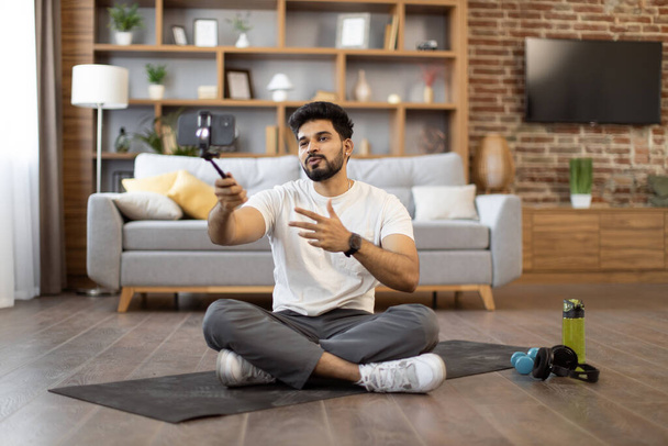 Active indian athlete in gym clothes holding tripod with smartphone and recording video content for sports blog. Attractive bearded male making online tutorial for followers via phone camera at home. - Foto, afbeelding