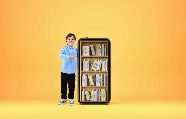 Kid smiling finger pointing at smartphone with digital library, books on rack, yellow background. Concept of e-learning and mobile app - Zdjęcie, obraz