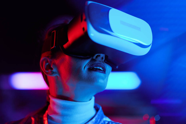 Neon metaverse futuristic concept. Portrait of modern middle aged woman in vr headset on dark background. - Foto, Imagem