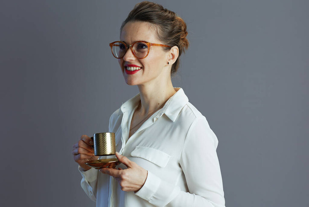 happy modern middle aged woman employee in white blouse with glasses and coffee cup isolated on grey background. - Foto, imagen