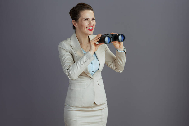 happy trendy middle aged business woman in a light business suit with binoculars isolated on gray background. - Foto, Imagen