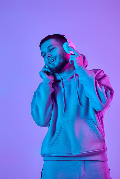 handsome bearded man with headphones in sweatshirt enjoying favorite music on purple neon background. Neon lighting - Фото, изображение
