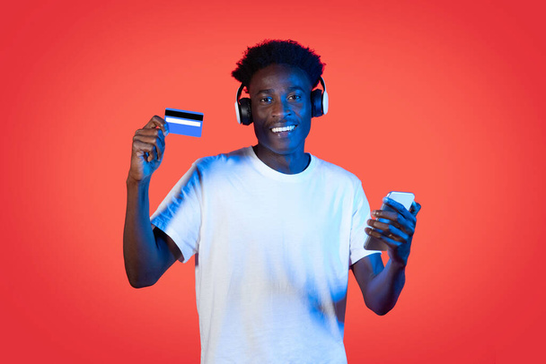 Cool vrolijke knappe jonge zwarte man hipster met draadloze hoofdtelefoon winkelen online op rode achtergrond in neon licht, met behulp van mobiele telefoon en bankkaart. Concept e-handel - Foto, afbeelding