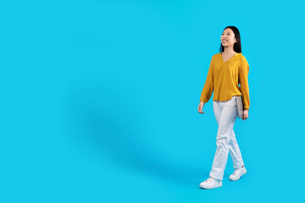 Feliz atraente jovem mulher asiática brilhante em roupa elegante nômade digital com laptop moderno caminhando em direção ao espaço de cópia sobre o fundo do estúdio azul, comprimento total. Freelance, conceito de trabalho remoto - Foto, Imagem