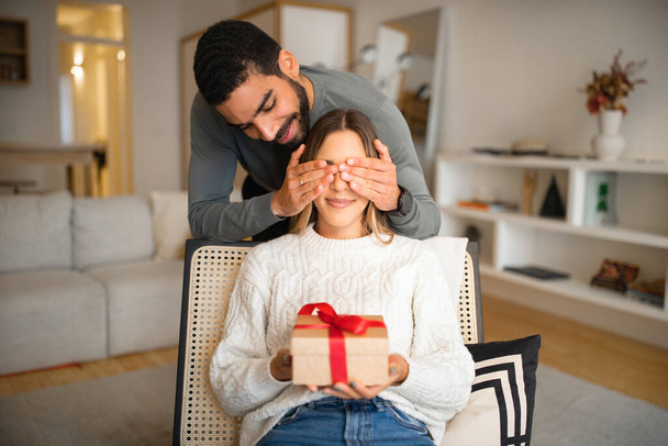 Cheerful multiethnic millennial family celebrating birthday, anniversary, holiday, husband close eyes to wife and give gift box in living room interior. Surprise, relationship and love at home - Photo, Image