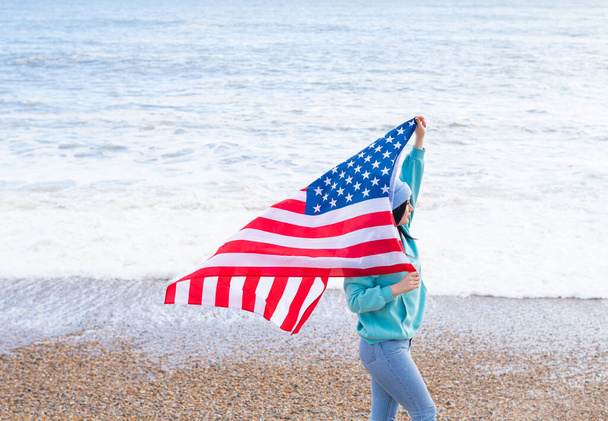 brunette woman in blue sweater and hat with american national flag near sea on the coastline - Photo, Image