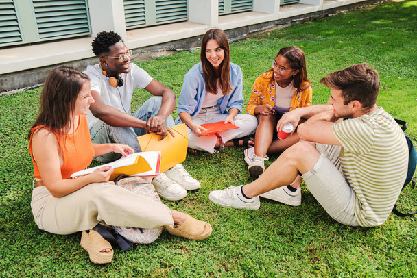 Grupa wielorasowych nastolatków siedzących na trawniku, rozmawiających i uśmiechających się razem na kampusie uniwersyteckim. Młodzi znajomi z college 'u udają się na sanki i uczą na trawie. Koncepcja edukacji. Wysoka jakość - Zdjęcie, obraz