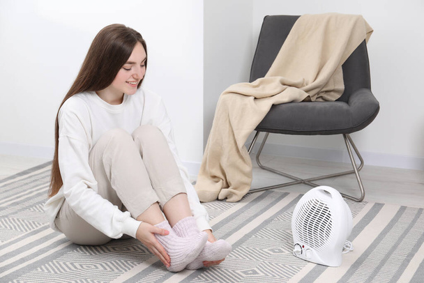 Young woman warming feet near electric fan heater at home - Foto, afbeelding