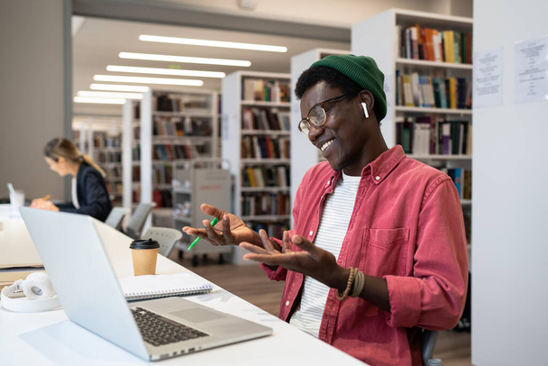 Agréable satisfait afro-américain gars étudiant ou freelancer gestes avec les mains dans la conversation à l'aide d'un ordinateur portable pour la vidéoconférence. Homme noir souriant assis à la bibliothèque en utilisant un appel vidéo pour l'éducation. - Photo, image