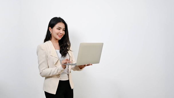 Beautiful millennial Asian businesswoman in a formal business suit using her laptop while standing against an isolated white background. - Fotó, kép