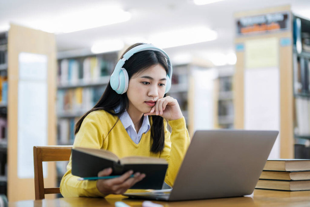 Young female college, highschool student wearing headphones and in casual clothings sitting at desk reading book, studying and doing research using laptop for school project at a library. E-Learning and Educational concept. - Fotó, kép