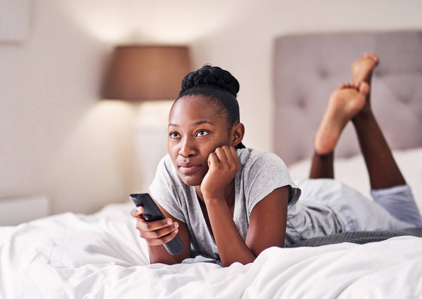 Aburrido de ver lo mismo de siempre. una joven sosteniendo un mando a distancia mientras está acostada en su cama - Foto, Imagen