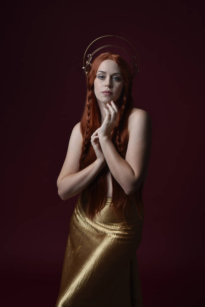 Lähikuva fantasia muotokuva kaunis nainen malli punaiset hiukset, jumalatar silkki kylpytakit & kulta kruunu. Poseeraus elehtivillä käsillä ojentaen, eristettynä tummanpunaisella studiotaustalla  - Valokuva, kuva