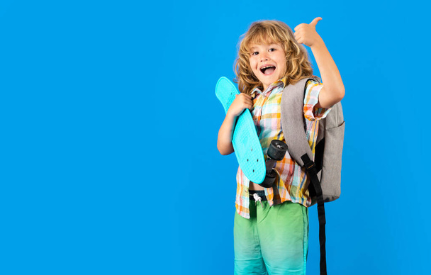 Grappig vrolijk kind met duim omhoog. Foto van kleine jongen houden skateboard geïsoleerd op blauwe achtergrond. Casual cultuur. Kind met skateboard en stijlvolle kleding in de studio - Foto, afbeelding