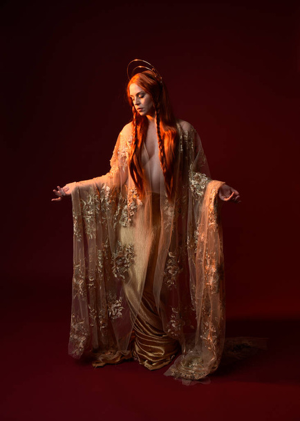 Full length fantasy portrait of beautiful woman model with red hair, goddess silk robes & gold crown. Standing pose gestural hands reaching out isolated on dark red studio background  - 写真・画像