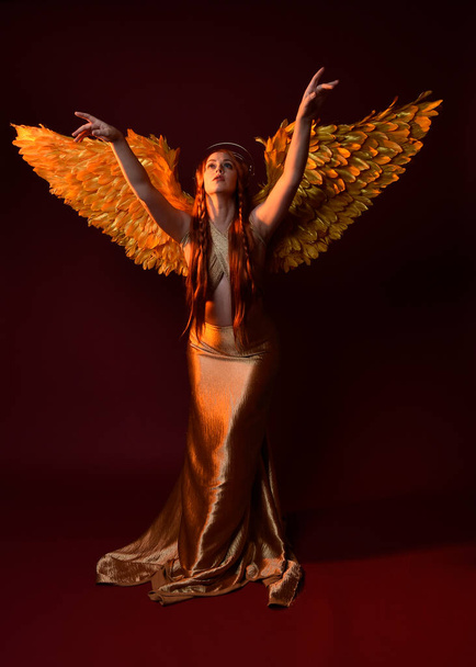 Full length πορτρέτο της όμορφης γυναίκας μοντέλο με μακριά κόκκινα μαλλιά, χρυσά μεταξωτά ρόμπες, στέμμα & φτερό φαντασίας φτερά αγγέλου. Μόνιμη στάση χειρονομιακή χέρια φθάνοντας απομονωμένο σε σκούρο κόκκινο φόντο στούντιο - Φωτογραφία, εικόνα