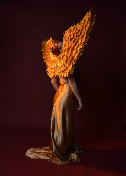 Full length portrait of beautiful woman model with long red hair, gold silk robes, crown & fantasy feather angel wings. Standing pose gestural hands reaching out isolated on dark red studio background - Φωτογραφία, εικόνα