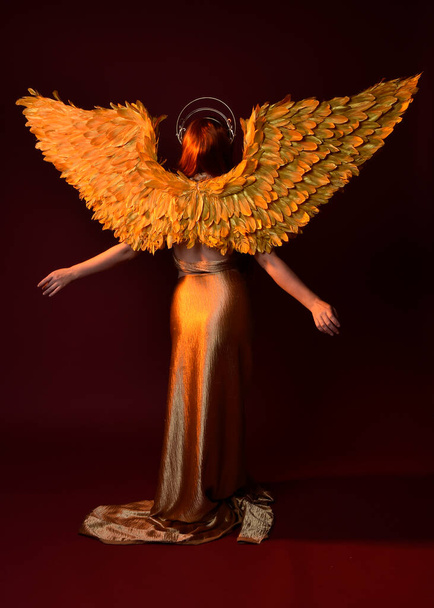 Full length portrait of beautiful woman model with long red hair, gold silk robes, crown & fantasy feather angel wings. Standing pose gestural hands reaching out isolated on dark red studio background - Photo, Image