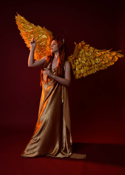 Full length portrait of beautiful woman model with long red hair, gold silk robes, crown & fantasy feather angel wings. Standing pose gestural hands reaching out isolated on dark red studio background - Photo, Image