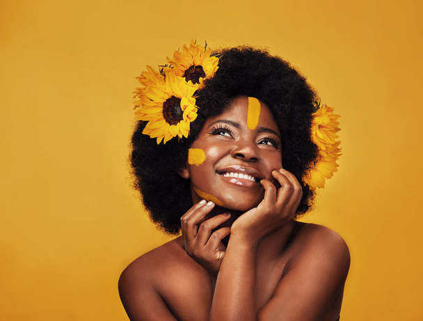 Such bliss can only blossom from within. Studio shot of a beautiful young woman smiling while posing with sunflowers in her hair against a mustard background - Photo, Image