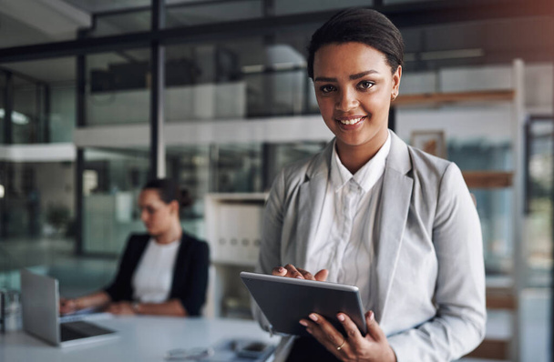 Im connected to everything in the business world. Portrait of an attractive young businesswoman using a digital tablet inside an office with her colleague in the background - Fotografie, Obrázek