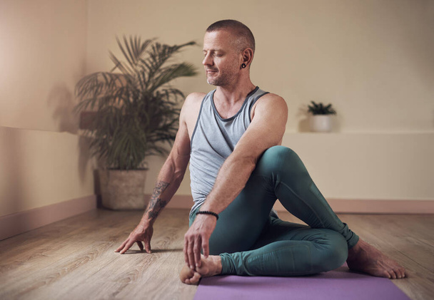Estirar la columna vertebral. Foto completa de un joven guapo sentado y sosteniendo un giro medio espinal durante una sesión de yoga en interiores - Foto, imagen