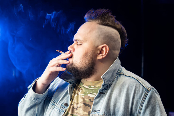 L'homme avec un mohawk sur la tête fume une cigarette blanche la nuit sur un fond noir dans la fumée. concept de tabagisme et mode de vie nocif. dépendance à la nicotine, dépendance à la nicotine - Photo, image