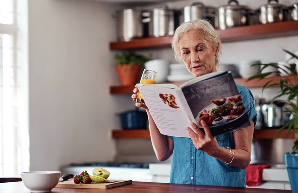 Iets speciaals maken uit het receptenboek. een aantrekkelijke oudere vrouw die leest uit een receptenboek tijdens het ontbijt in de keuken thuis - Foto, afbeelding