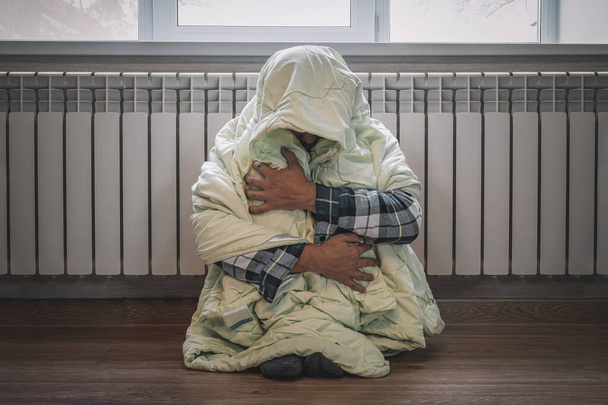В зимнее время дома холодно. Человек замерзает в своем доме зимой из-за сломанного термостата. Грустный молодой парень, завернутый в клетчатую дрожь, сидя на полу в гостиной - Фото, изображение