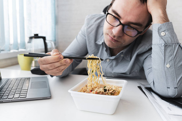 Νέος δυσαρεστημένος άνθρωπος τρώει στιγμιαία noodles ενώ εργάζεται με φορητό υπολογιστή στο γραφείο. Γεύμα στο γραφείο. άγευστο πρόχειρο φαγητό - Φωτογραφία, εικόνα