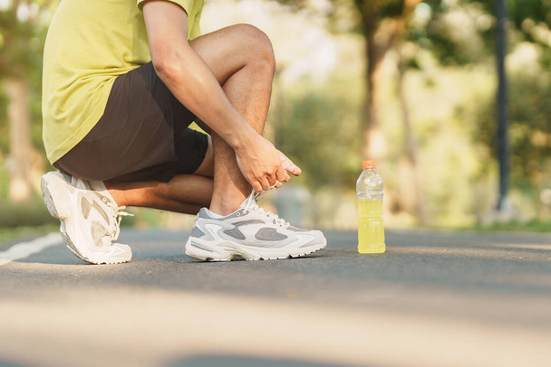 Young athlete man tying running shoes with Energy Drink water, male runner ready for jogging outside, asian Fitness walking and exercise in the park morning. wellness, wellbeing and sport concepts - Photo, Image