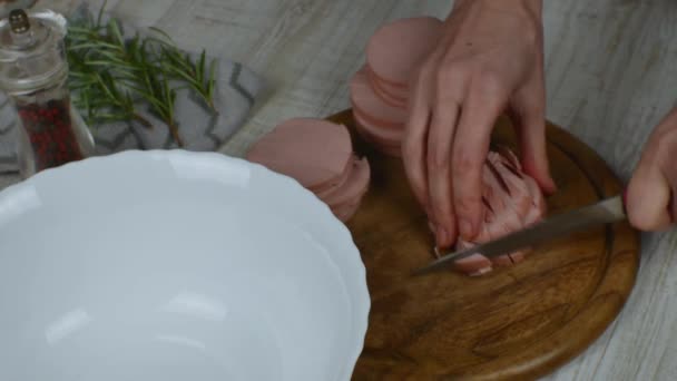 Sliced boiled sausage slices are laid out on a wooden cutting board and cut into small pieces to prepare a meat salad. Pepper grinder, sprig of rosemary next to cutting board. Womens hands close up. - Footage, Video
