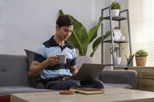 Beau millénial homme buvant du café et naviguant sur Internet sur ordinateur portable dans le salon confortable.  - Photo, image