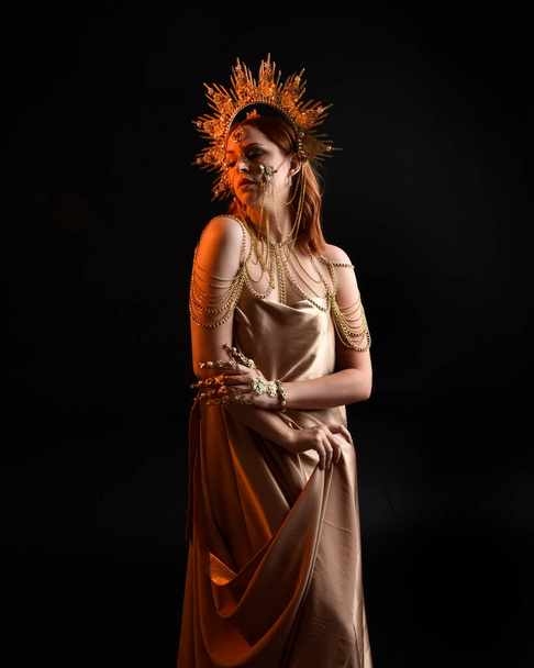 Gros plan portrait fantaisie de belle femme modèle aux cheveux roux, robes de soie déesse & couronne ornée d'or. Poser avec les mains gestuelles tendues vers l'extérieur, isolé sur fond sombre studio  - Photo, image