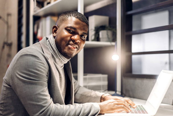 Jonge zwarte Afrikaanse man ontspannen met behulp van technologie van laptop computer tijdens het zitten op tafel.Jonge creatieve Afrikaanse man werken en typen op toetsenbord thuis. - Foto, afbeelding