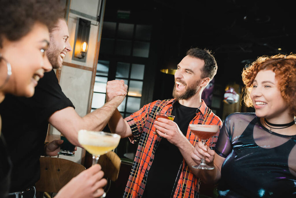 Positive men holding hands near happy interracial friends with cocktails in bar  - Photo, Image