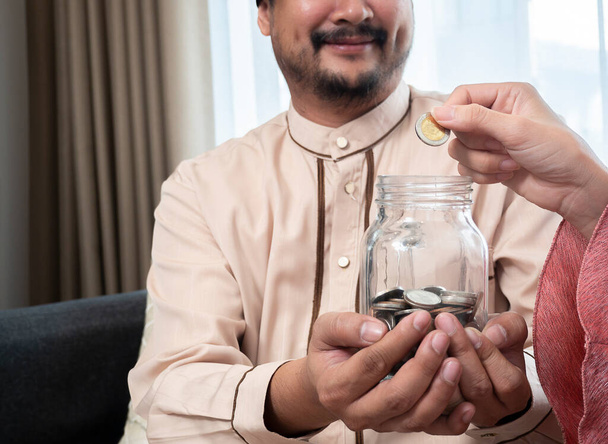 Cropped image of a happy couple at home saving money in a jar glass. Close up hands of a happy Asian muslim couple at home saving money in a jar glass and smiling - Foto, afbeelding