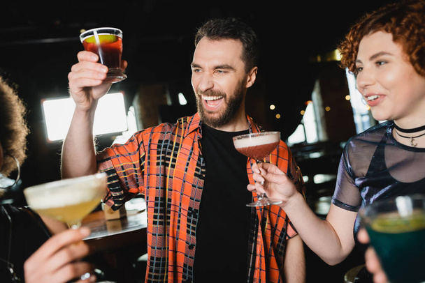 Cheerful bearded man holding negroni cocktail near friends with glasses in bar  - Photo, Image