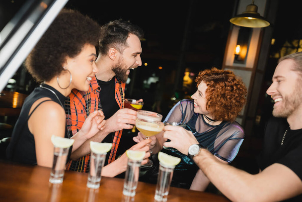 Seitenansicht multiethnischer Freunde mit verschiedenen Cocktails, die sich in der Bar in der Nähe verschwommener Tequila-Gläser unterhalten  - Foto, Bild