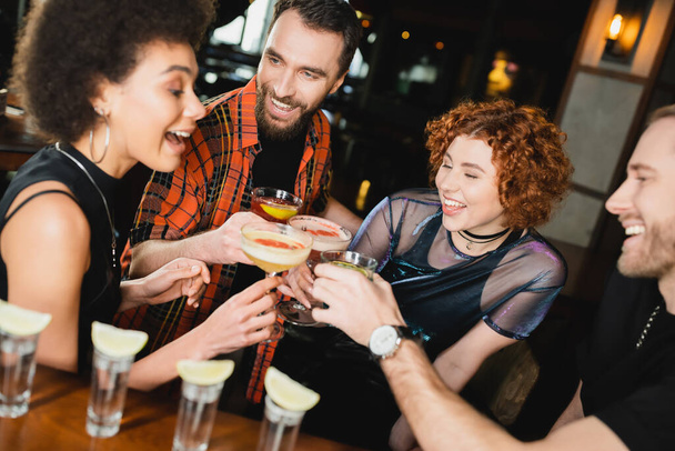 Fröhliche multiethnische Menschen klappern in der Nähe von Tequila-Shots in einer Bar verschiedene Cocktails  - Foto, Bild